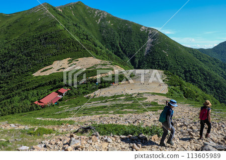 Two female climbers walking along the mountain trail of Mt. Jonen with Jonen hut and Mt. Yokotsutsu 113605989