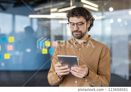 A focused business professional stands in a modern office, using a tablet. His casual business attire and the office's glass background with sticky notes add to the corporate atmosphere. A focused business professional stands in a modern office, using a tablet. His casual business attire and the office's glass background with sticky notes add to the corporate atmosphere. 113608081
