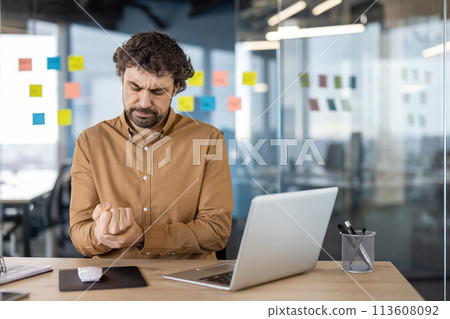 Male office worker experiencing discomfort, clutching his hand in pain at his workplace with laptop and stationary around. 113608092