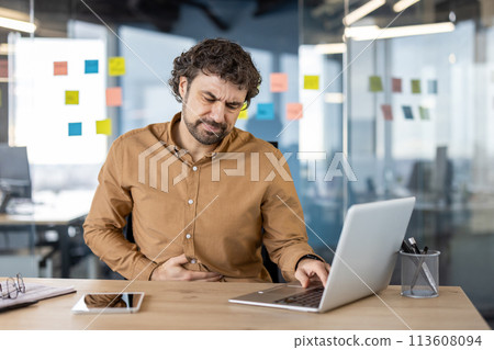 Adult male office worker feeling discomfort, experiencing a painful stomachache while working on a laptop in a modern office setting. 113608094