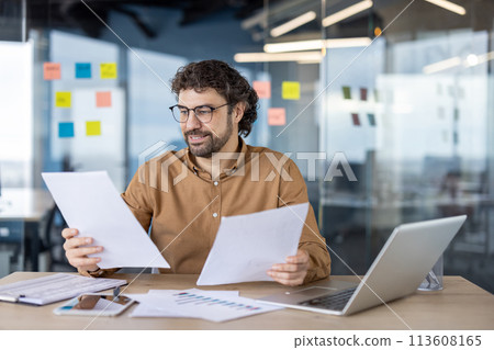 A mature Hispanic man smiling while analyzing papers with a laptop in a modern office setting. A mature Hispanic man smiling while analyzing papers with a laptop in a modern office setting. 113608165