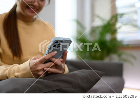 Cropped shot of young woman sitting on couch at home and typing message on mobile phone 113608299
