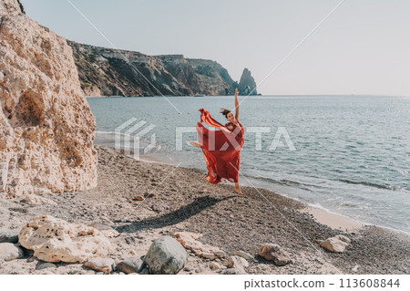 Woman red dress sea. Female dancer in a long red dress posing on a beach with rocks on sunny day. Girl on the nature on blue sky background. Woman red dress sea. Female dancer in a long red dress posing on a beach with rocks on sunny day. Girl on the nature on blue sky background. 113608844
