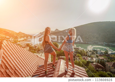 women standing on rooftop, enjoys town view and sea mountains. Peaceful rooftop relaxation. Below her, there is a town with several boats visible in the water. Rooftop vantage point. 113608928