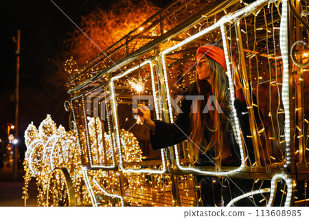Woman holding sparkler night while celebrating Christmas outside. Dressed in a fur coat and a red headband. Blurred christmas decorations in the background. Selective focus Woman holding sparkler night while celebrating Christmas outside. Dressed in a fur coat and a red headband. Blurred christmas decorations in the background. Selective focus 113608985