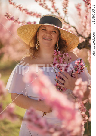 Woman blooming peach orchard. Against the backdrop of a picturesque peach orchard, a woman in a long white dress and hat enjoys a peaceful walk in the park, surrounded by the beauty of nature. Woman blooming peach orchard. Against the backdrop of a picturesque peach orchard, a woman in a long white dress and hat enjoys a peaceful walk in the park, surrounded by the beauty of nature. 113609015