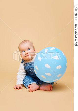 Cheerful infant, cute baby-girl in classic overalls laying with blue balloon against beige color studio background. Cheerful infant, cute baby-girl in classic overalls laying with blue balloon against beige color studio background. 113610885