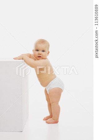 Curious baby peeks around cube, leaning on it for support, showcasing tearly childhood against white studio backdrop. 113610889