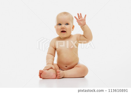 Little, curious baby raises tiny hand in gentle wave, sitting bare-skinned except for diaper against white studio background. 113610891