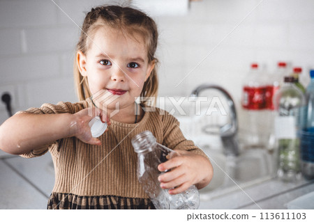 A child removes the cap from a plastic bottle for recycling and looks expressively at the camera. Beautiful girl child helps sort plastic in the kitchen of her home. Plastic recycling concept. A child removes the cap from a plastic bottle for recycling and looks expressively at the camera. Beautiful girl child helps sort plastic in the kitchen of her home. Plastic recycling concept. 113611103