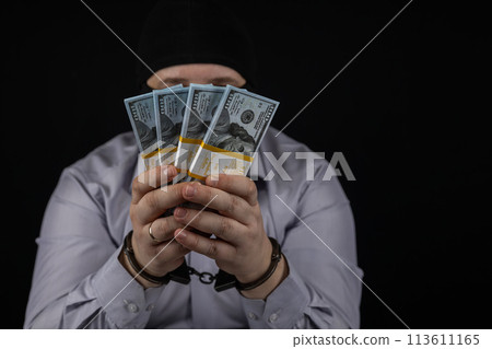 A man holds in his hands a large amount of money for which he was arrested. A man in a balaclava and handcuffs holds dollars on a black background. A man holds in his hands a large amount of money for which he was arrested. A man in a balaclava and handcuffs holds dollars on a black background. 113611165