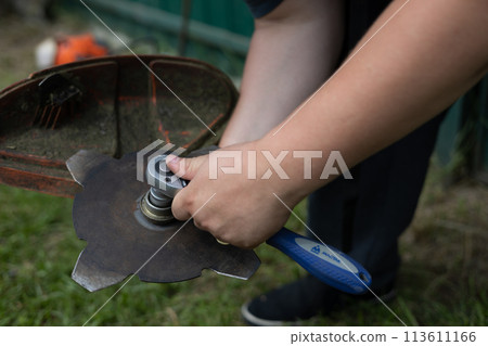 A man uses a wrench to fix the grass cutting attachment on a trimmer. Close-up. In the summer, a man installs a grass trimmer attachment in his yard. Grass trimmer concept. A man uses a wrench to fix the grass cutting attachment on a trimmer. Close-up. In the summer, a man installs a grass trimmer attachment in his yard. Grass trimmer concept. 113611166