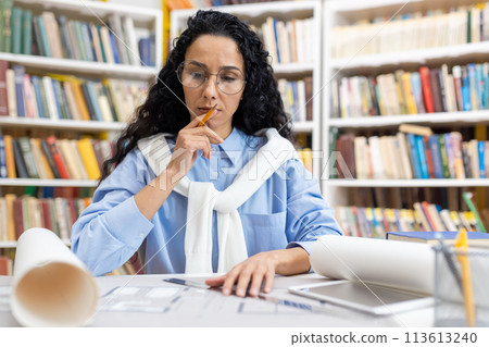 A professional female architect deeply engrossed in examining building blueprints at a library desk. Busy and thoughtful atmosphere with books in the background. A professional female architect deeply engrossed in examining building blueprints at a library desk. Busy and thoughtful atmosphere with books in the background. 113613240