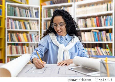 Focused female architect reviewing blueprints at a library table, with a positive expression and books in the background. Focused female architect reviewing blueprints at a library table, with a positive expression and books in the background. 113613241