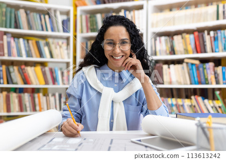 Cheerful female architect examining building plans in a library setting, smiling joyfully with bookshelves in the background. 113613242