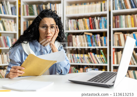 A distressed woman with curly hair and glasses reads a troubling letter from a yellow envelope at her desk, surrounded by bookshelves. A distressed woman with curly hair and glasses reads a troubling letter from a yellow envelope at her desk, surrounded by bookshelves. 113613253