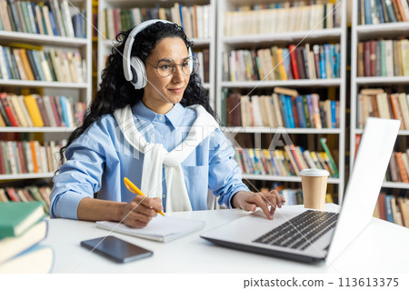 A female student wearing headphones concentrates on her laptop while studying in a library. She is surrounded by books and has coffee nearby, indicating a long study session. 113613375