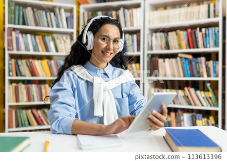 A cheerful woman wearing headphones is comfortably using a digital tablet in a well-stocked library, surrounded by numerous books. A cheerful woman wearing headphones is comfortably using a digital tablet in a well-stocked library, surrounded by numerous books. 113613396