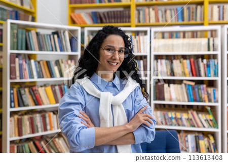 Portrait of a confident, smiling female librarian standing with arms crossed in a brightly colored library full of books. She wears glasses and a casual outfit. Portrait of a confident, smiling female librarian standing with arms crossed in a brightly colored library full of books. She wears glasses and a casual outfit. 113613408