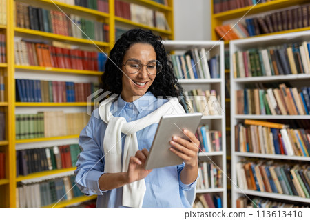 Smiling young woman in glasses using a digital tablet among bookshelves in a library, representing modern learning and technology in education. 113613410