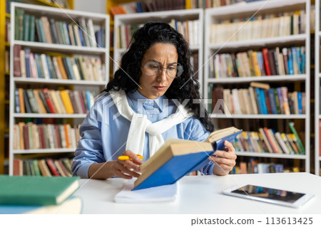 A dedicated student engrossed in reading a book surrounded by a vast collection of literature in a well-stocked library. A dedicated student engrossed in reading a book surrounded by a vast collection of literature in a well-stocked library. 113613425