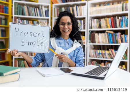 Cheerful female teacher holding a board with 'Present Simple' written on it, demonstrating English grammar in a library setting surrounded by books. 113613429