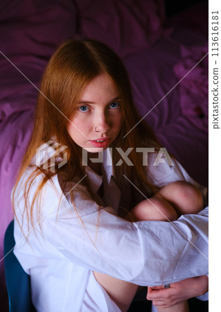 Amazing ginger woman in long white shirt sitting at the chair Fashion portrait Studio shot Beautiful red hairs girl in white at home on the pink backgtound bed hugs the legs with her hands 113616181