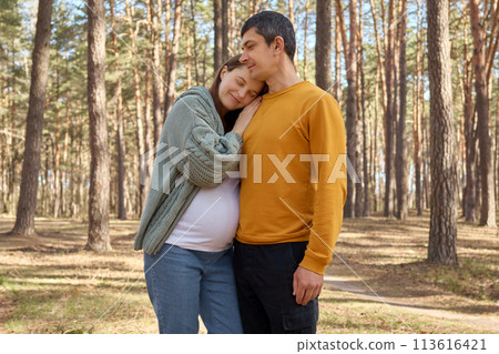 Beautiful young family walking in spring forest hugging and admiring beautiful forest pregnant woman leaning on husband shoulder enjoying happy moments 113616421
