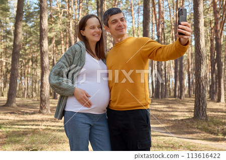 Loving man and pregnant woman walking in park hugging couple making selfie in spring forest husband holding smartphone capturing happy moments of walking during pregnancy Loving man and pregnant woman walking in park hugging couple making selfie in spring forest husband holding smartphone capturing happy moments of walking during pregnancy 113616422
