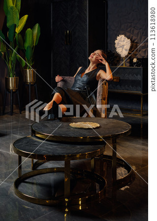 Beautiful young woman in leather dress sits in armchair near coffee table 113618980