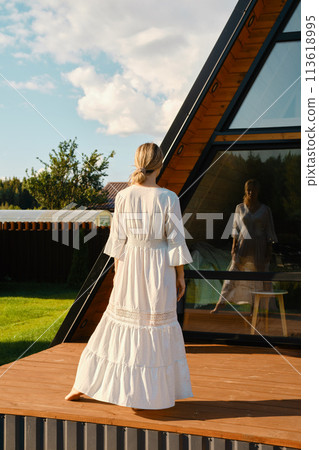 Barefoot woman standing on wooden terrace of tiny house and looking at her reflection in the window Barefoot woman standing on wooden terrace of tiny house and looking at her reflection in the window 113618995