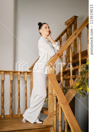 A pensive woman stands on a flight of stairs with her elbow resting on the staircase railing 113619015