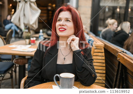 Beautiful woman with bright red lipstick sitting on restaurant terrace. Wonderful woman with red hair in black clothes posing in cafe. Beautiful woman with bright red lipstick sitting on restaurant terrace. Wonderful woman with red hair in black clothes posing in cafe. 113619952