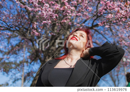 Red haired woman wearing stylish outfit near blossoming sakura in park. Fashionable spring look. Springtime blooming female portrait. Red haired woman wearing stylish outfit near blossoming sakura in park. Fashionable spring look. Springtime blooming female portrait. 113619976