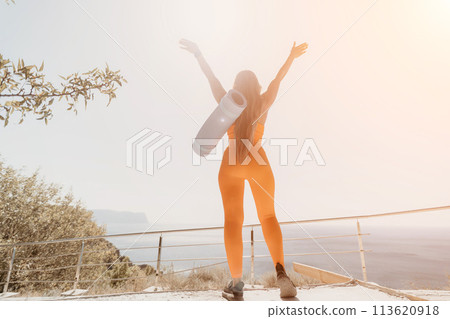 Fitness woman sea. Happy middle aged woman in orange sportswear exercises morning outdoors on yoga mat with laptop in park over ocean beach. Female fitness pilates yoga routine. Healthy lifestyle. 113620918