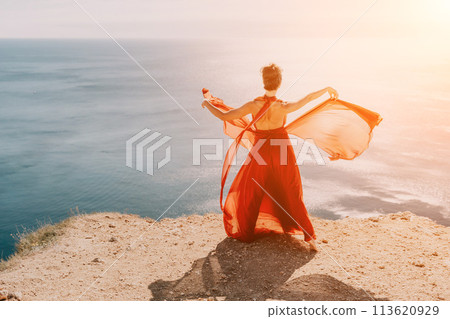 Woman in red dress on sea. Side view a Young beautiful sensual woman in a red long dress posing on a rock high above the sea on sunset. Girl on the nature on blue sky background. Fashion photo. Woman in red dress on sea. Side view a Young beautiful sensual woman in a red long dress posing on a rock high above the sea on sunset. Girl on the nature on blue sky background. Fashion photo. 113620929