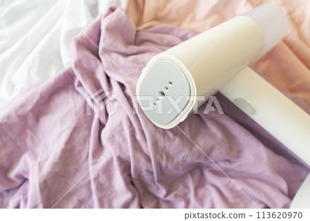 Handheld steamer rests on crumpled purple and white fabrics, ideal for quickly removing wrinkles and caring for clothes. 113620970