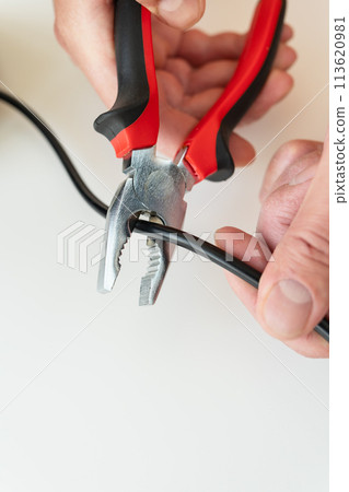 Close-up of hands using red and black pliers to cut a black cable on a white background. Close-up of hands using red and black pliers to cut a black cable on a white background. 113620981