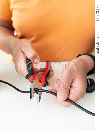 A person in an orange shirt cutting a black electrical cord with red and black handled pliers. A person in an orange shirt cutting a black electrical cord with red and black handled pliers. 113620982