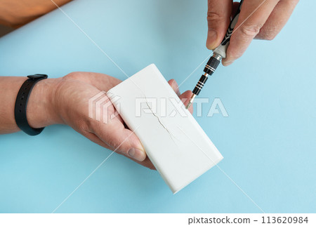 Hands skillfully repair a white power bank with a soldering iron on a blue background. Hands skillfully repair a white power bank with a soldering iron on a blue background. 113620984