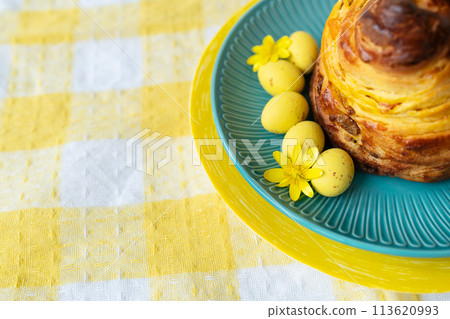 A delightful pastry on a teal plate with yellow Easter eggs and a flower, set on a checkered tablecloth. A delightful pastry on a teal plate with yellow Easter eggs and a flower, set on a checkered tablecloth. 113620993