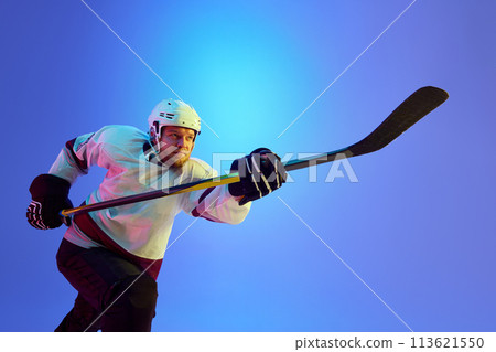 Ambitious man, hockey player posing with stick, showing his determination against gradient blue background in neon light 113621550