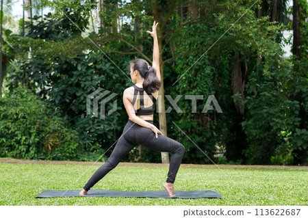 Asian woman practicing yoga in Warrior Pose (Virabhadrasana) on the mat in outdoor park. Asian woman practicing yoga in Warrior Pose (Virabhadrasana) on the mat in outdoor park. 113622687