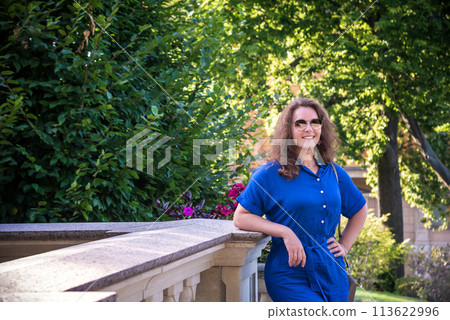 Beautiful young smiling woman in sunglasses in city park at summer day wearing dress and looking to the camera Beautiful young smiling woman in sunglasses in city park at summer day wearing dress and looking to the camera 113622996