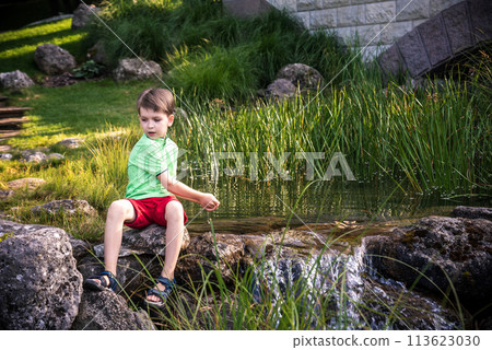 A young boy sits on the river bank, dreams. Warm summer or spring day. Cute Caucasian kid play with water 113623030