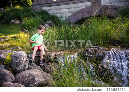 A young boy sits on the river bank, dreams. Warm summer or spring day. Cute Caucasian kid play with water 113623031