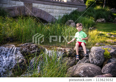 A young boy sits on the river bank, dreams. Warm summer or spring day. Cute Caucasian kid play with water 113623032