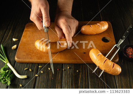The chef uses a knife to cut a Vienna sausage on a kitchen board. Grill cooking concept with sausages and aromatic spices. American cuisine The chef uses a knife to cut a Vienna sausage on a kitchen board. Grill cooking concept with sausages and aromatic spices. American cuisine 113623383