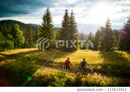 Two cyclists men riding electric bikes outdoors at sunset. Back view of male tourists resting on the hill, enjoying beautiful mountain landscape in the evening. Concept of active leisure. 113623912