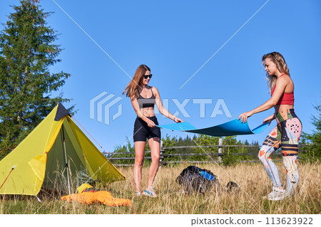 Two women hikers camping in mountains. Female tourists making campsite in hills, holding mat, enjoying nature. Concept of tourism and traveling, Two women hikers camping in mountains. Female tourists making campsite in hills, holding mat, enjoying nature. Concept of tourism and traveling, 113623922